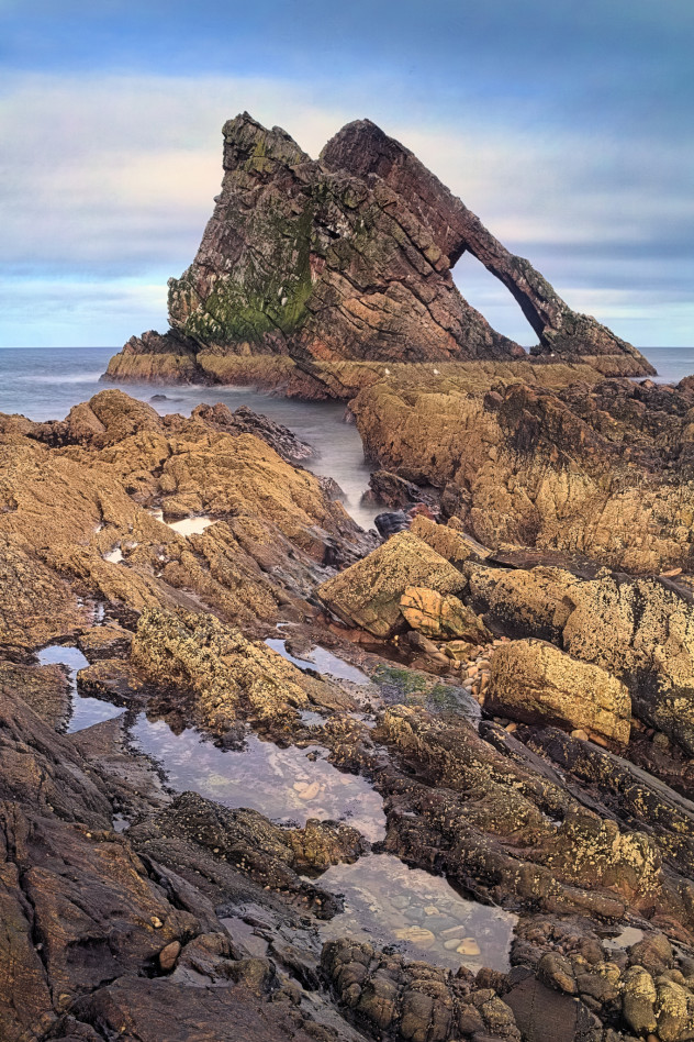 A classic view - the strong angular shape of Bow Fiddle with rock-pools in the foreground, Portknockie.

This was a real blighter of a shot to make. Even with the tripod legs not very extended and wedged into crevices in the rockpool, the wind was such that even a few seconds' exposure was impossible. Added to this, the light seen here was so transient, it blossomed to optimum golden-yellow on the stone and vanished again within the course of about 10 seconds.